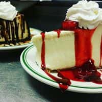 Two slices of cheesecake, one in the front topped with strawberry syrup and whip cream, and one in the background topped with chocolate syrup and whipped cream