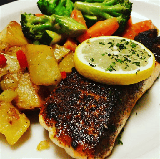 charred fish topped with a slice of lemon, mixed vegetables and home fries.