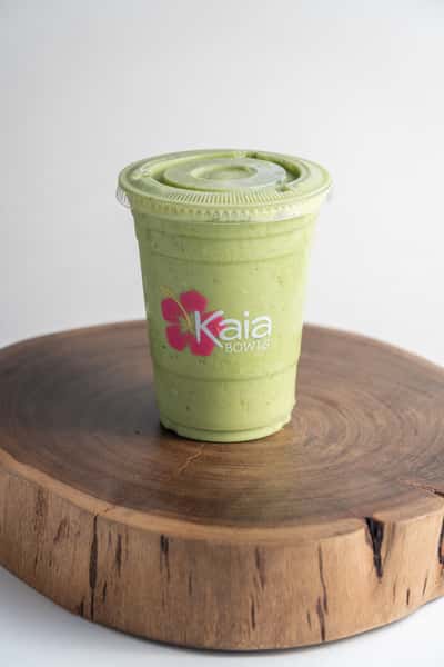 A light green smoothie in a clear plastic cup with a pink hibiscus logo sits on a rustic wood slice against a plain white background.