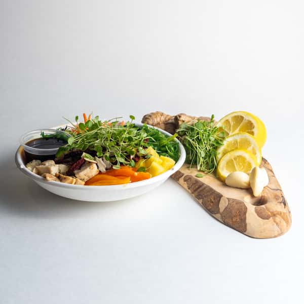 A vibrant healthy bowl with chicken, microgreens, and fresh vegetables, presented with ginger, lemon, and garlic on a wooden board.