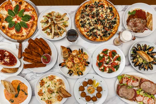 top-down spread of assorted pizzas, pasta, and appetizers arranged on a table for a family-style meal