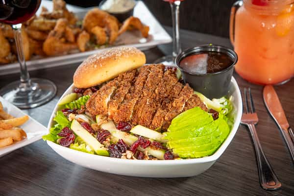 NinaFresh salad topped with crispy fried chicken, avocado slices, dried cranberries, apple strips, and a side of dark dressing, served with a soft sesame bun