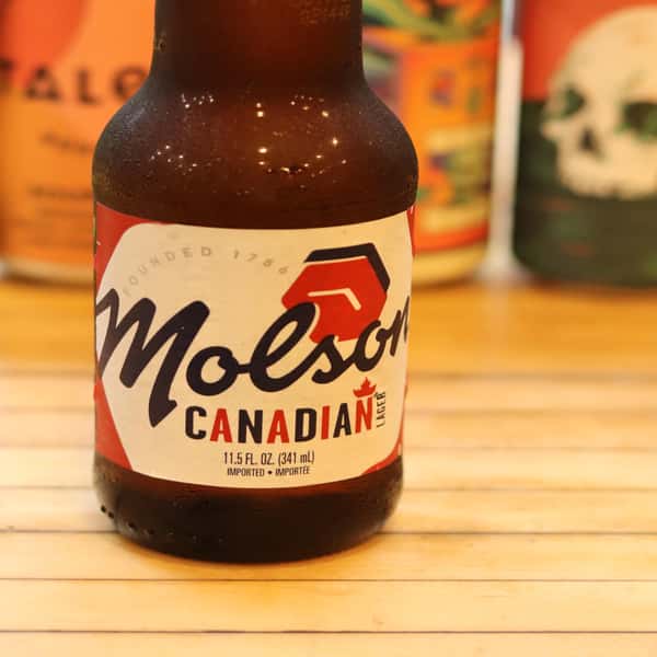 A bottle of Molson Canadian Lager with a clear label displaying "Founded 1786" and the iconic Molson logo, with other cans in the background.