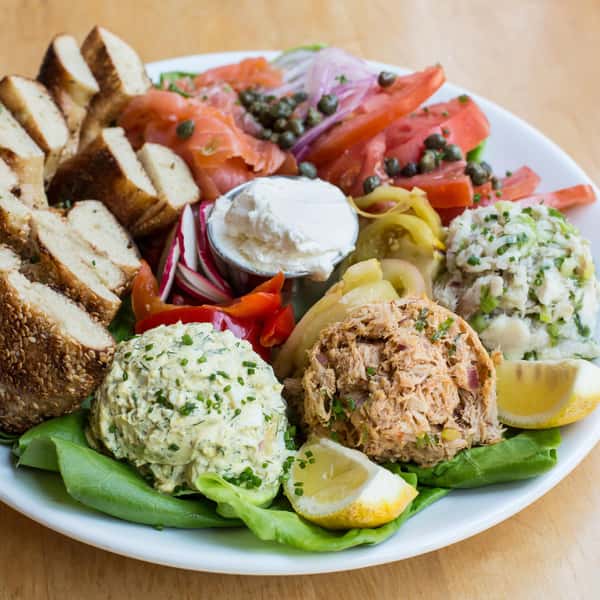 A platter with assorted smoked salmon, sliced bagels, pickled vegetables, cream cheese, and three different salads: egg salad, tuna salad, and potato salad, garnished with lemon wedges.