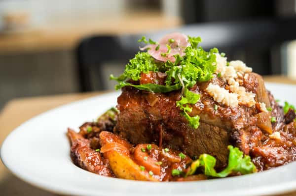 A close-up shot of a plated beef roast or brisket, with tender potatoes, topped with greens, pickled onions, and a sprinkling of herbs or cheese. The dish is drenched in a savory sauce.