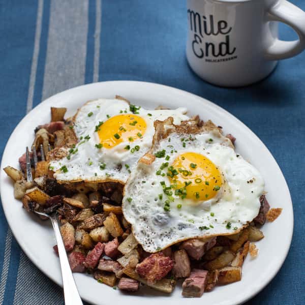 A plate of crispy hash browns and diced meats topped with two sunny-side-up eggs, garnished with chives, with a Mile End coffee mug in the background.