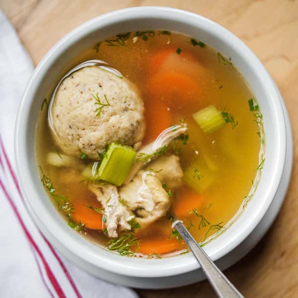 A bowl of chicken soup with a matzo ball, carrots, celery, and herbs, served with a spoon on the side.