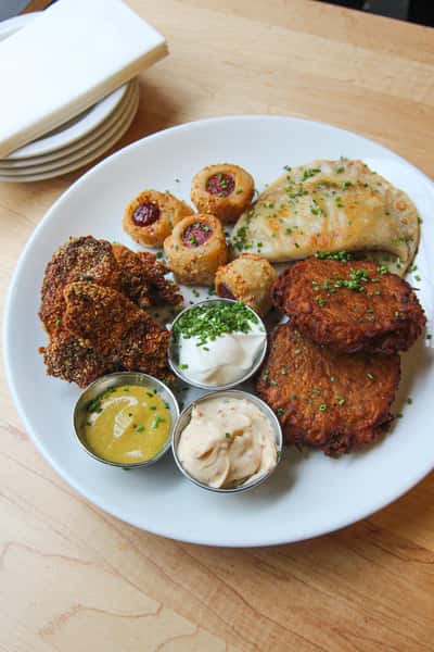 A plate with crispy latkes, fried chicken, potato knishes, a stuffed pierogi, and sauces including mustard, sour cream, and a creamy dip.