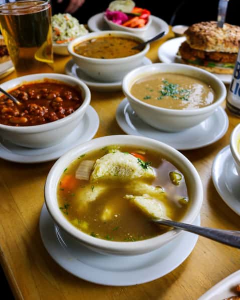 A variety of soups including chicken with matzo balls, chili, and creamy soup, served with a side of coleslaw, pickled vegetables, and a sandwich.