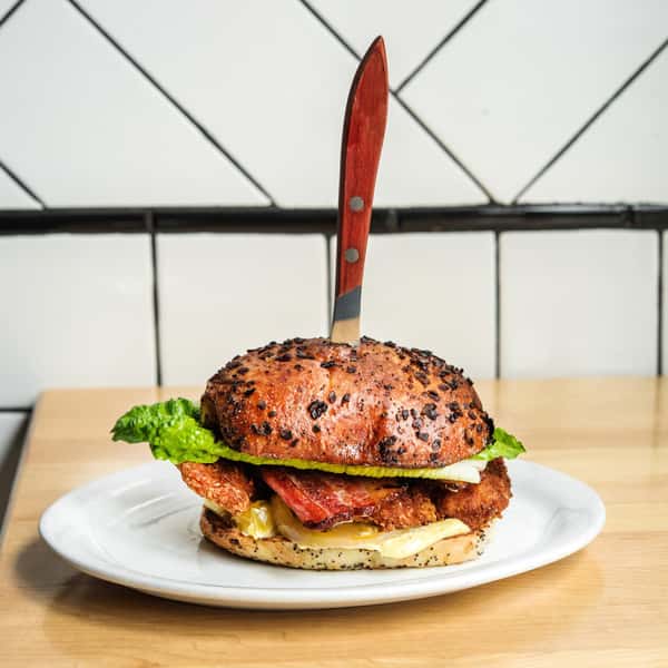 A sandwich with crispy bacon, lettuce, and other toppings, served on a poppy seed bun with a knife inserted through the top, placed on a white plate.