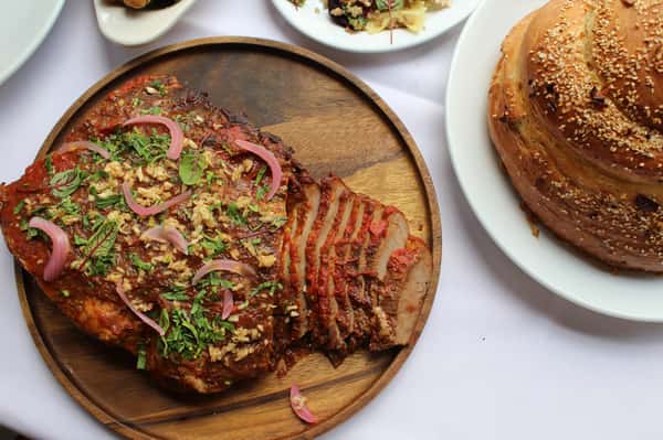 A platter of sliced roast beef with a tangy sauce, garnished with pickled onions, fresh herbs, and served with sesame bread.