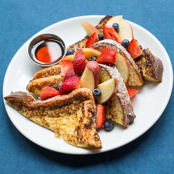 A plate of French toast topped with fresh strawberries, blueberries, and apple slices, dusted with powdered sugar, and served with a side of syrup.