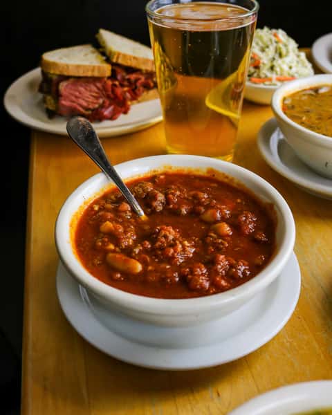 A bowl of hearty chili with beans and meat, accompanied by a glass of beer, coleslaw, and a deli sandwich with layers of roast beef.