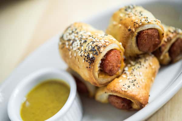 A plate of savory pig-in-a-blanket, with mini sausages wrapped in sesame seed and poppy seed-studded pastry, served with a side of mustard for dipping.