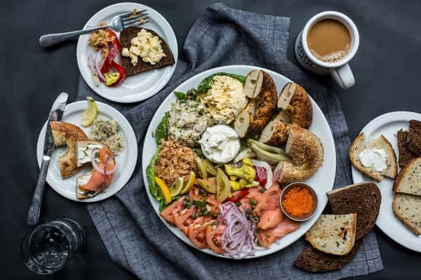 A plate of bagels with smoked salmon, tomatoes, onions, pickled vegetables, and cream cheese, alongside a cup of coffee.