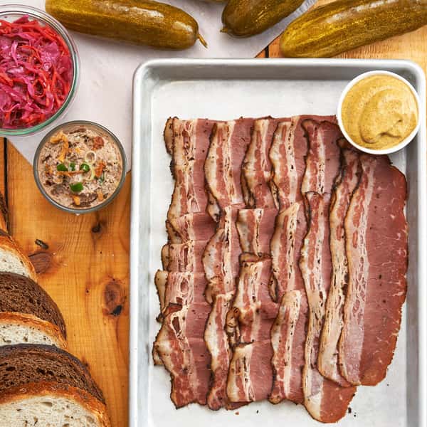 A platter of sliced pastrami, mustard, pickles, pickled cabbage, and rye bread slices, served on a tray.