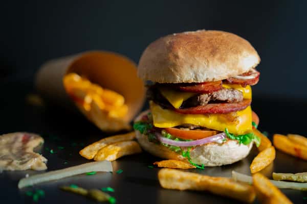 A cheeseburger topped with bacon and vegetables stands next to a container of fries at Spot Gourmet Burgers.