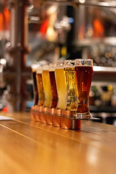 A row of six different beers in glasses, each with varying shades, placed on a wooden bar counter.