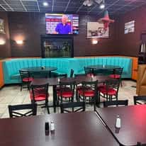 Restaurant interior with teal booth seating, black chairs with red cushions, and a TV mounted on a dark brown wall.
