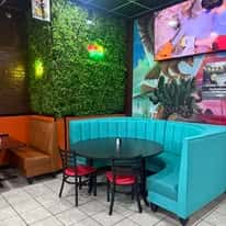 Colorful restaurant corner with a turquoise booth, round black table, red-cushioned chairs, green faux plant wall, and mural artwork.