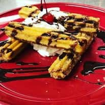 Churros drizzled with chocolate sauce, topped with whipped cream and a cherry on a red plate.