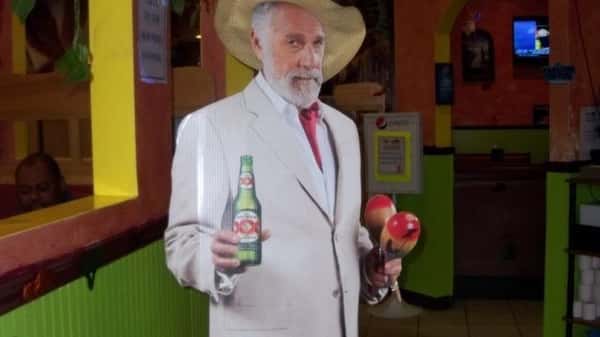 A life-size cutout of a man in a suit and sombrero, holding a bottle of beer and a pair of maracas, set in a brightly colored restaurant.