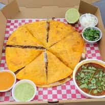 A quesadilla cut into slices, accompanied by lime, onions, cilantro, and two different sauces in small containers, with a side of stew.