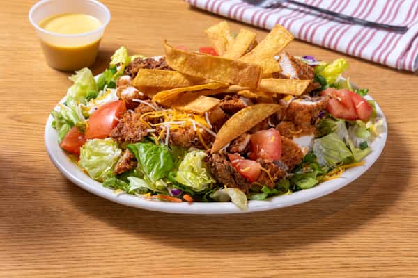 Southern fried chicken salad with crispy breaded chicken, fresh lettuce, tomatoes, shredded cheese, crunchy tortilla strips, and house dressing served in a white bowl.