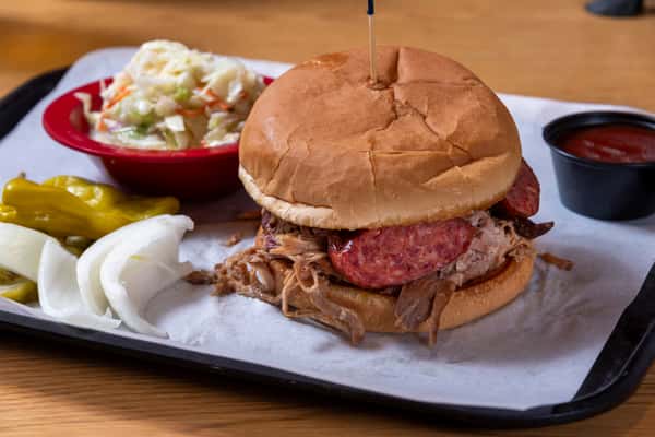 Pulled pork sandwich topped with sliced smoked sausage on a soft bun, served with coleslaw, pickles, onions, and BBQ sauce