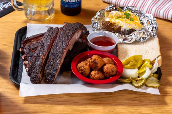 Smoked pork ribs served with a foil-wrapped loaded baked potato, hushpuppies, sliced white bread, pickles, and BBQ sauce