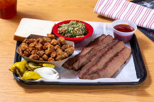 Sliced smoked brisket served with fried okra, herb salad, pickles, white bread, and barbecue sauce.