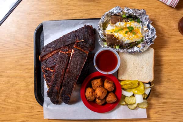 Southern BBQ rib plate with smoked pork ribs, hushpuppies, loaded baked potato, sliced white bread, and pickles.