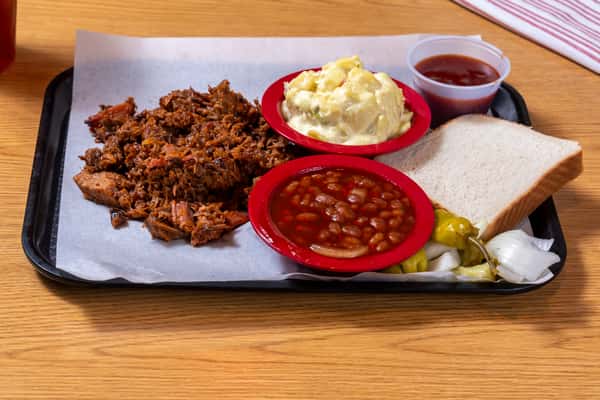 Smoked pulled pork served with baked beans, potato salad, white bread, pickles, onions, and barbecue sauce.