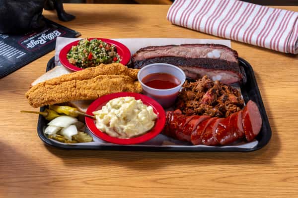 Southern BBQ combo platter featuring smoked brisket, pulled pork, sliced sausage, fried catfish, potato salad, baked beans, and barbecue sauce.