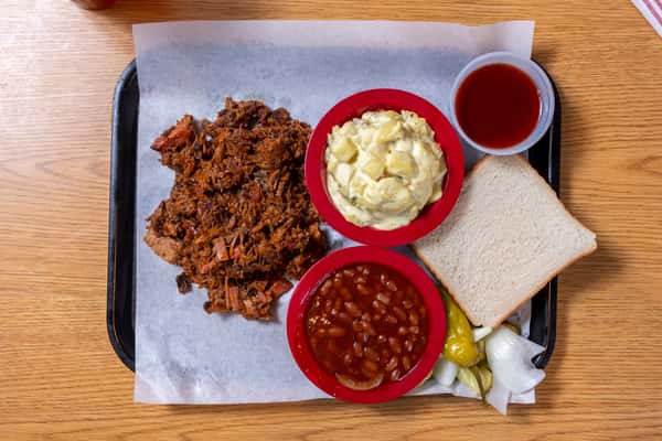 Slow-smoked pulled pork served with baked beans, creamy potato salad, white bread, pickles, onions, and barbecue sauce.