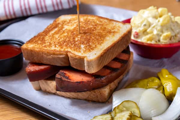 Grilled smoked sausage sandwich on toasted white bread with potato salad, pickles, and Southern barbecue sides.