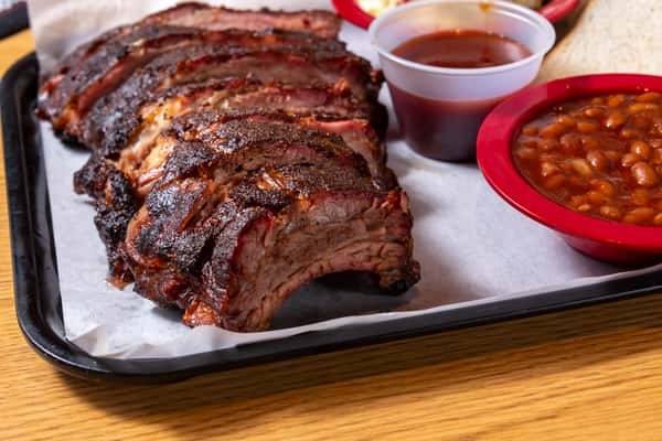Close-up of smoked pork ribs with dark bark served alongside baked beans and barbecue sauce.