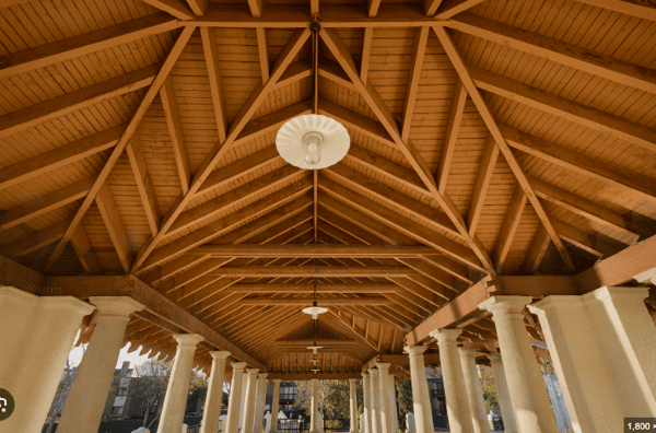 This image showcases the interior roof structure of an elegant open-air pavilion. The roof features a beautifully crafted wooden truss design with exposed beams and symmetrical support. Hanging pendant lights line the center, supported by a series of tall, white columns. The architectural style and materials suggest a historic or coastal setting, ideal for formal gatherings or events.