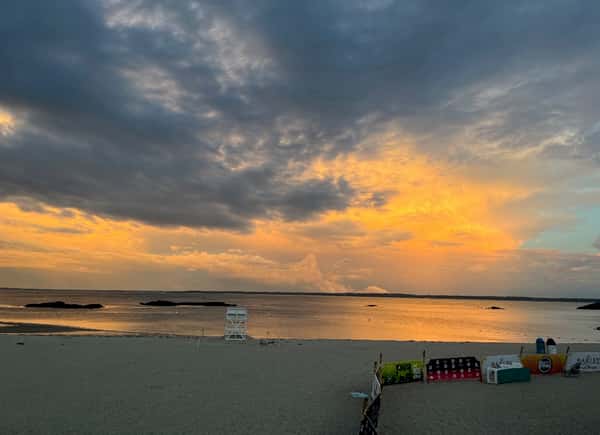 Dramatic sunset over a calm beach with golden clouds, a lifeguard chair, and scattered event barriers near the shoreline.