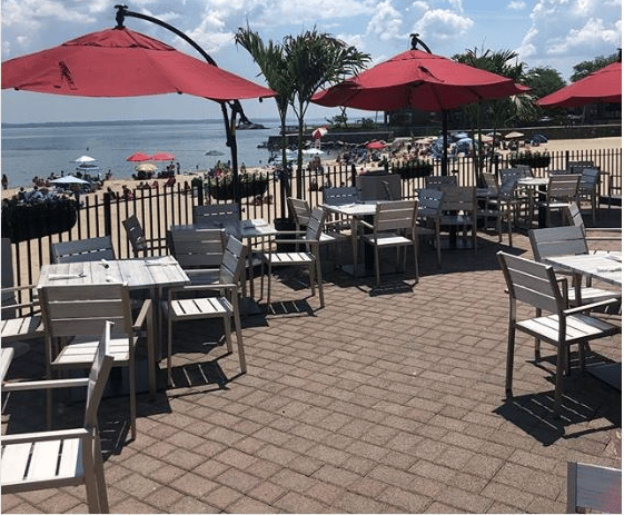 Outdoor beachfront patio with white tables and chairs, red umbrellas, and palm trees overlooking a sunny beach with colorful umbrellas.