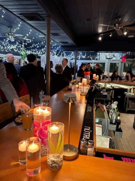 Elegant indoor bar setting with floating candles in glass vases, guests mingling beneath string lights during an evening event.
