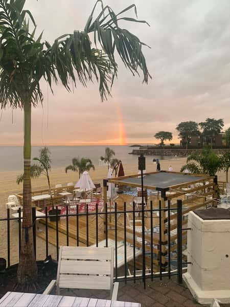 Palm trees and patio seating overlook a beach deck setup with tents at sunset, with a vibrant rainbow visible over the ocean horizon.