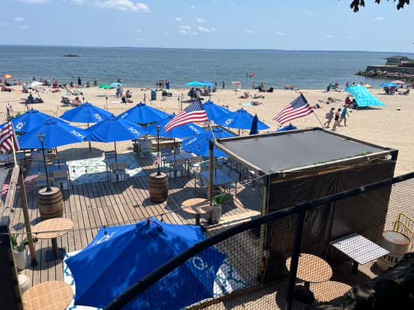 Wooden beach deck with blue umbrellas, American flags, and lounge seating, set against a backdrop of sunbathers and ocean waves.