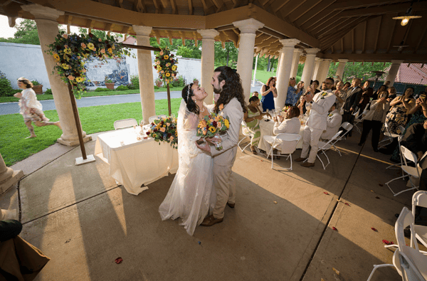 Bride and groom share a joyful dance under a gazebo surrounded by guests and floral arches during a sunny wedding reception.