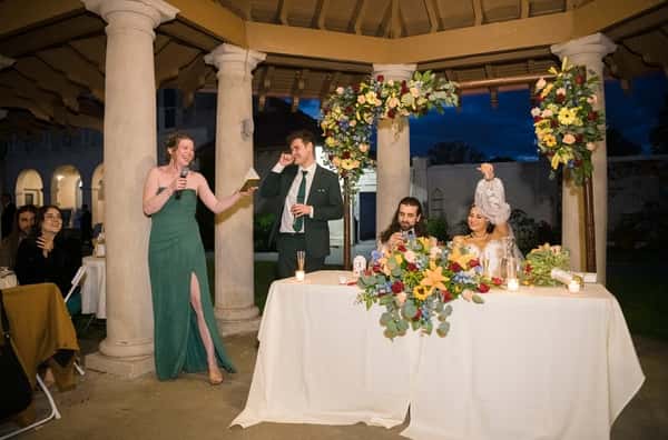 Wedding reception toast under a columned gazebo at night, with floral arrangements, a bride and groom at the table, and guests laughing.