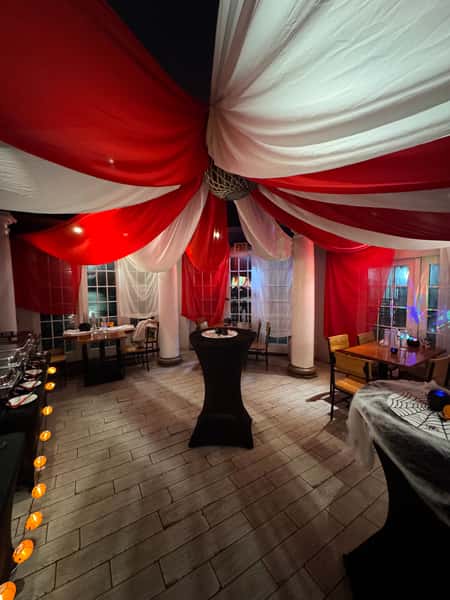 This image depicts an indoor space decorated with a dramatic red and white circus-style ceiling draping. The fabric is gathered at the center, forming a tent-like canopy over the room. The setting includes tables and chairs arranged for dining, with festive Halloween-themed decorations like cobwebs and orange lights shaped like pumpkins. The ambiance is moody and theatrical, perfect for a themed party or Halloween event.