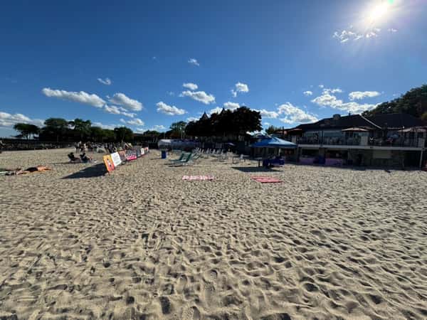 Sunny beach with scattered towels, lounge chairs, umbrellas, and people relaxing near a beachfront building under a bright blue sky.
