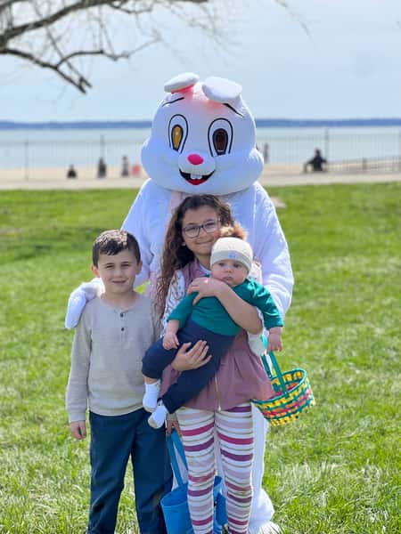 Three children pose with a person in an Easter Bunny costume on a grassy lawn near a beach with water in the background.