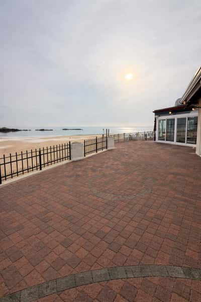 Brick patio overlooking a sandy beach and calm ocean, with a black fence and glass-walled venue on the right under a hazy sky.