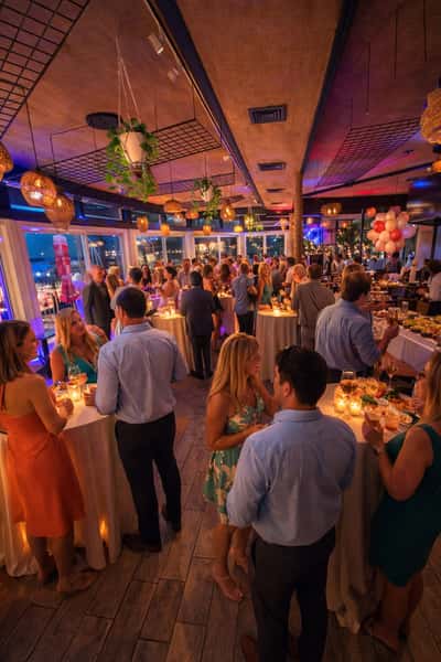 Elegant indoor evening party with guests mingling around candle-lit cocktail tables under warm lighting and hanging plants.